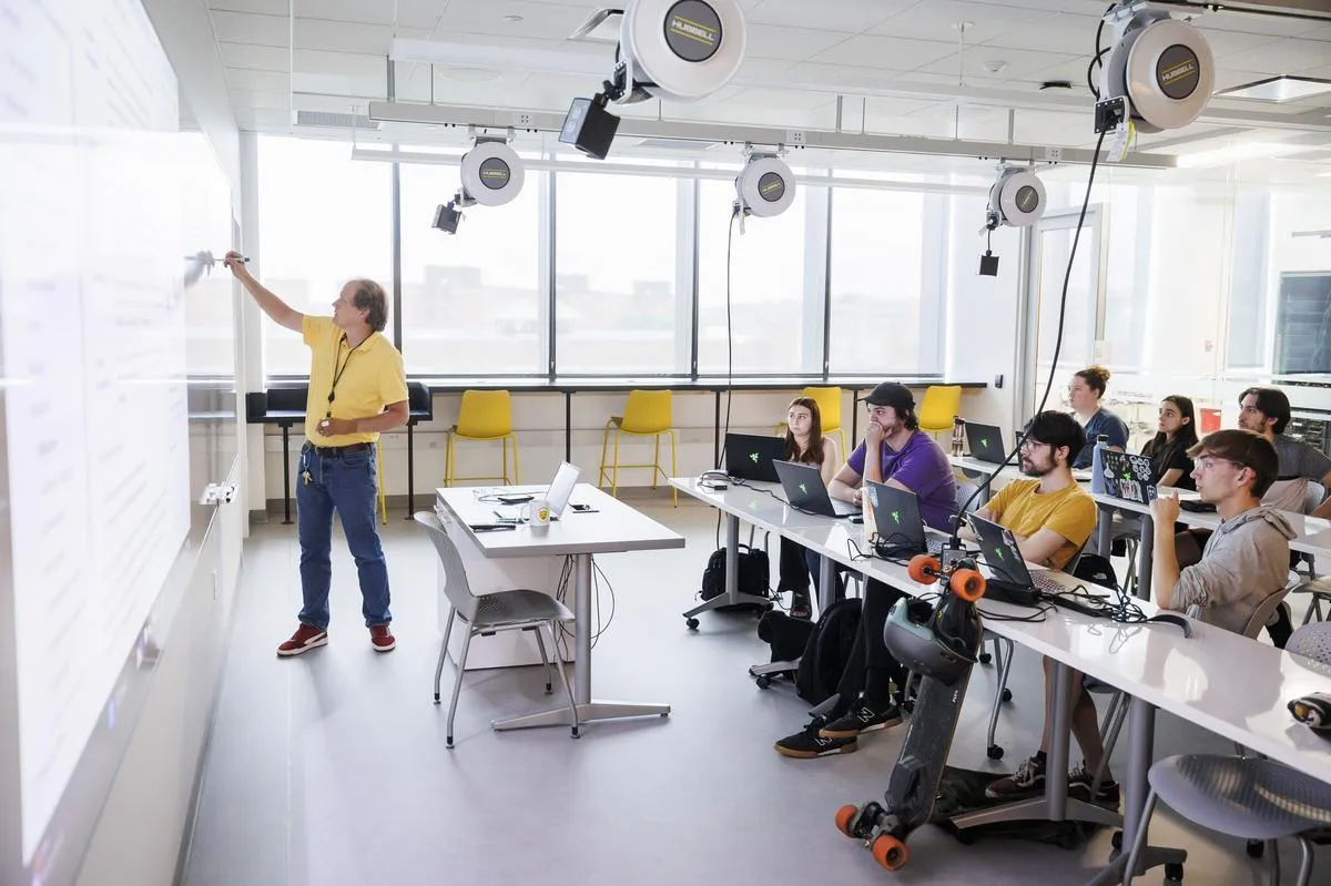 A faculty member draws on a white board in front of a class of students with their laptops.