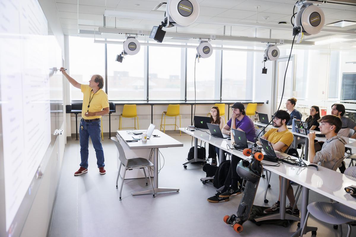 A faculty member draws on a white board in front of a class of students with their laptops.