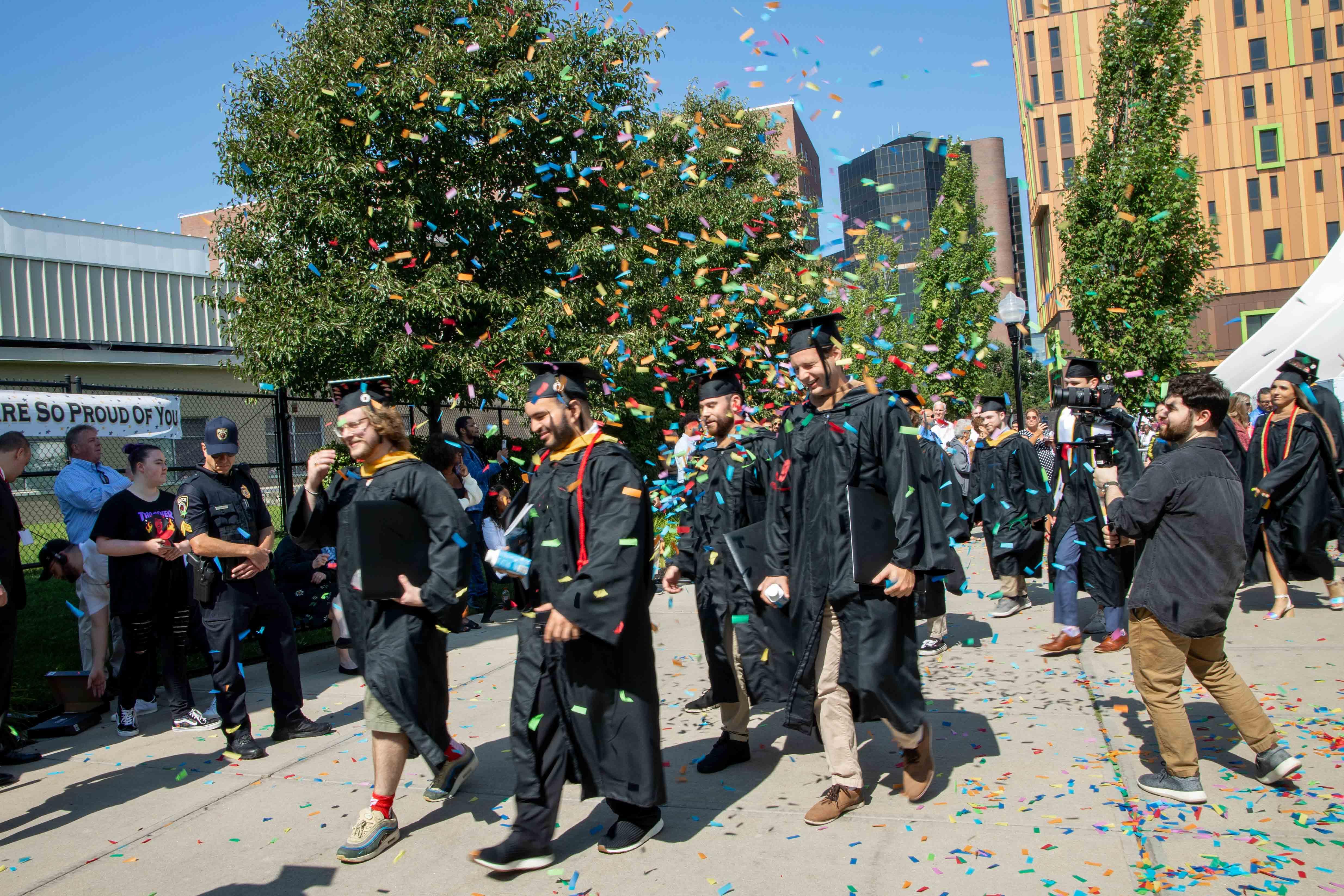 Students wearing black caps and gowns being showered with red, green, yellow confetti on a sunny day with blue sky in the background after their commencement ceremony