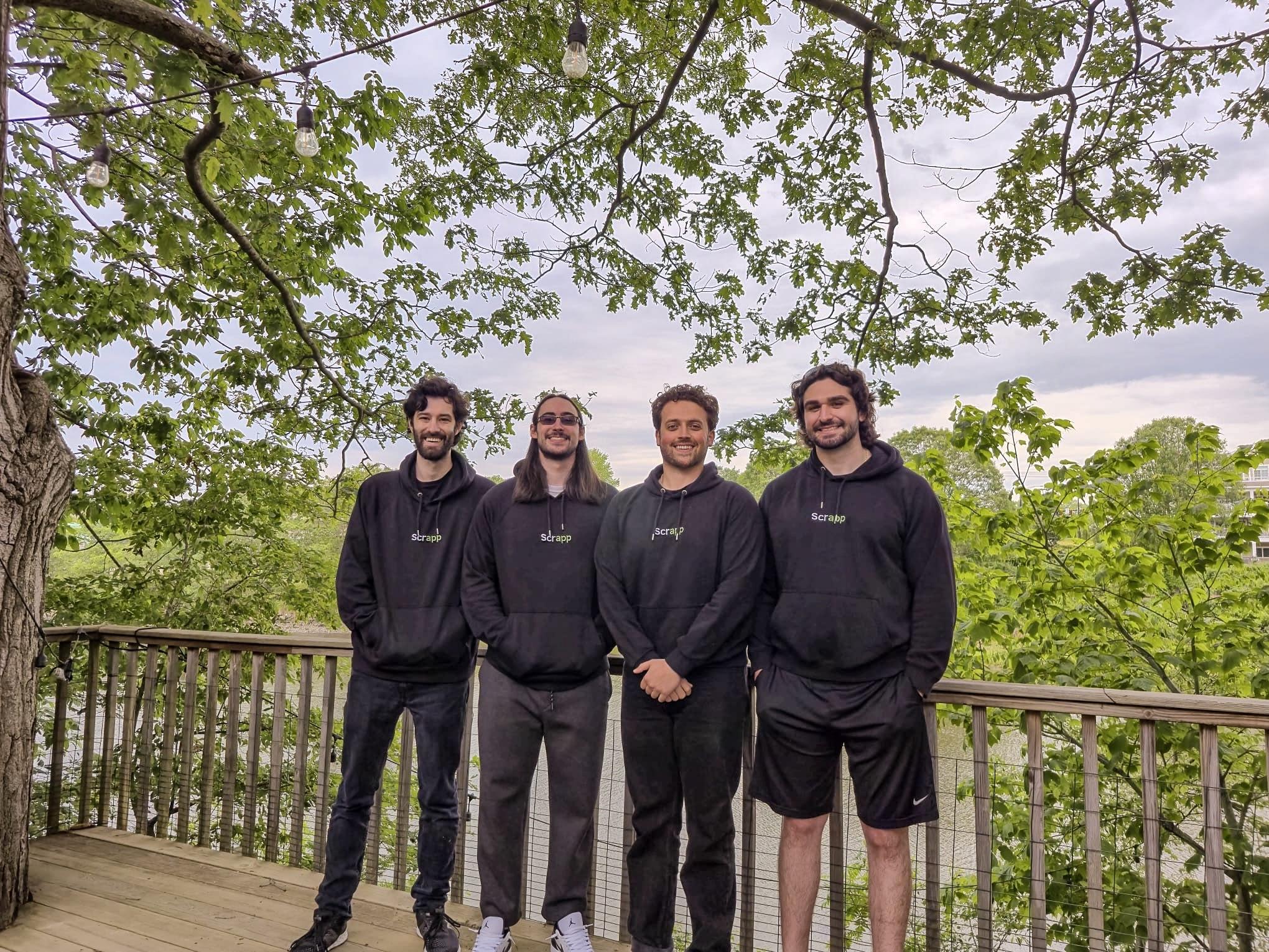 a group of four people pose in black sweatshirts in front of trees