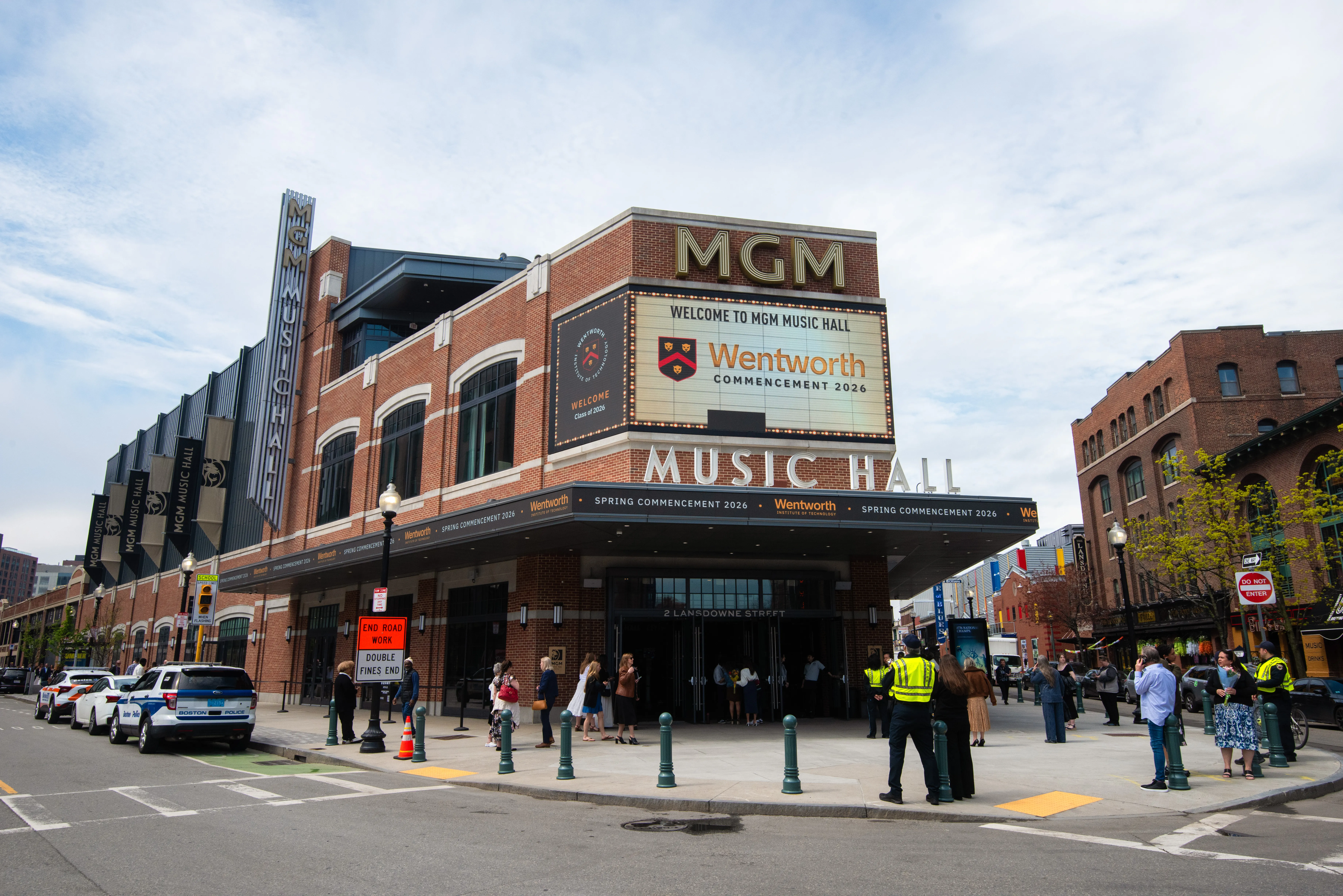 Marquee at MGM Music Hall where commencement is now held - brick building - corner view with cars lining the left including a Boston Police Car and view of the right side oof the street with families waiting to go in - several trees in bloom.