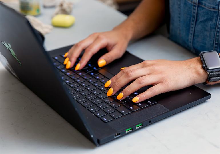 A woman's hands with yellow nail polish typing on a laptop.