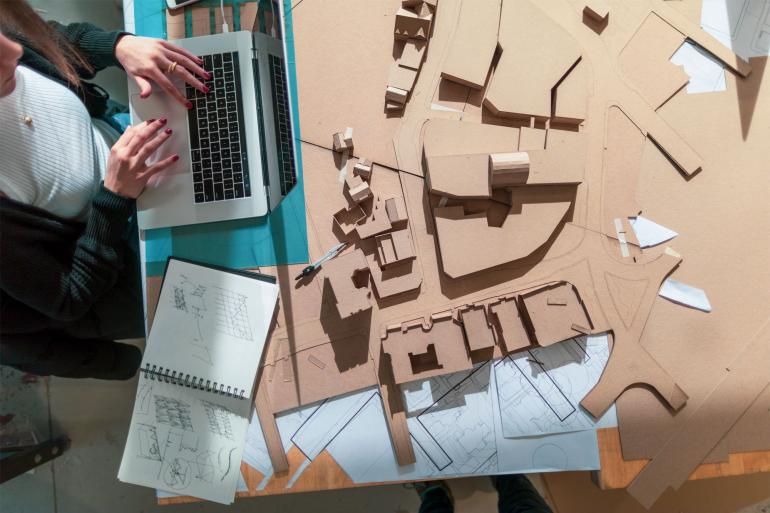 A woman's hands are in view as she works on her laptop surrounded by a cardboard architectural model.