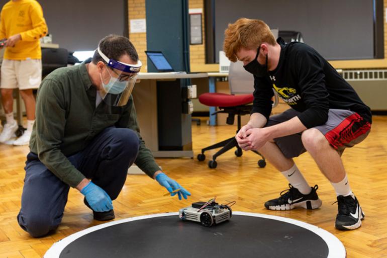 A professor and student kneel to examine a small car they designed on a track.