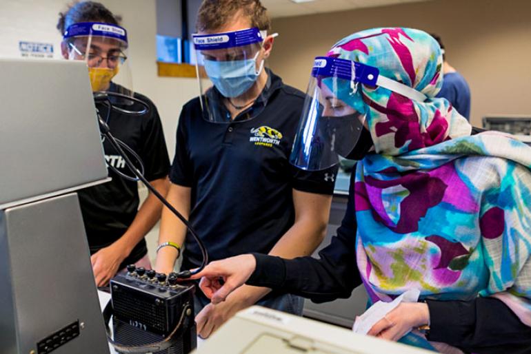 A female student in a color hijab works on a project with 2 male students.