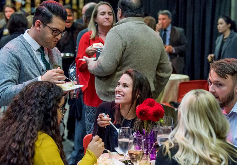 A group of faculty and staff, seated and standing, talk and eat at an event.