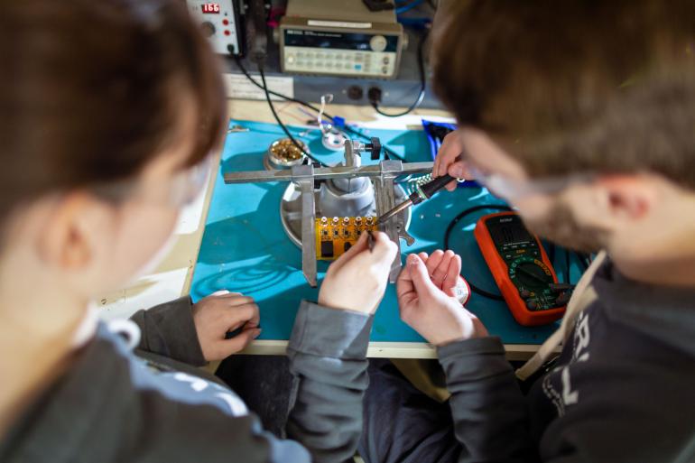 Two students are pictured from behind while they work together on an electrical device. 