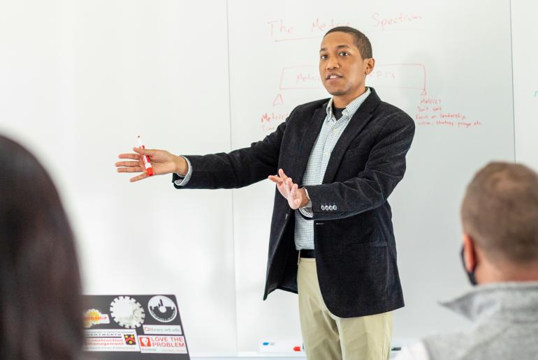 A young man stands in front of a white board addressing a classroom. 