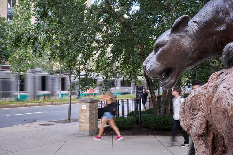 A woman walks past the leopard statue while the T passes in the background. 