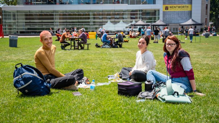 Three students sit on the quad and enjoy at barbecue on a sunny day