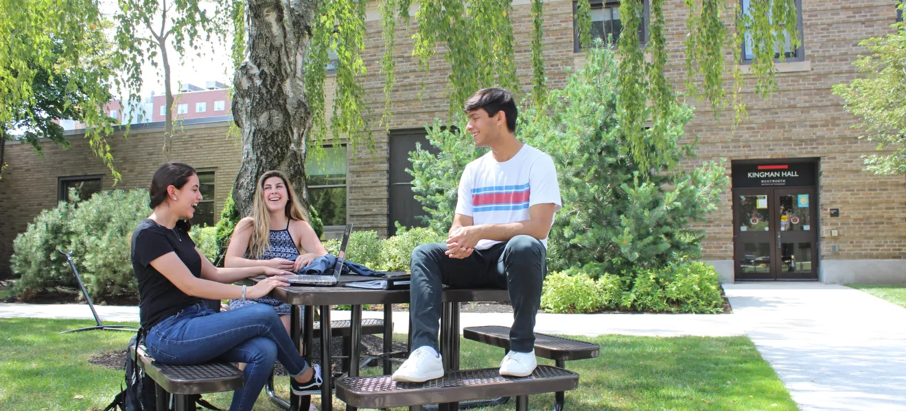 Three students sit at a picnic table in the quad while talking and smiling at one another