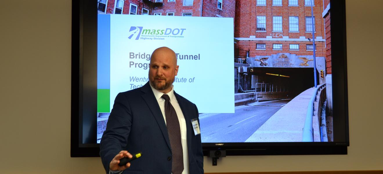 man speaking in front of a screen displaying a bridge