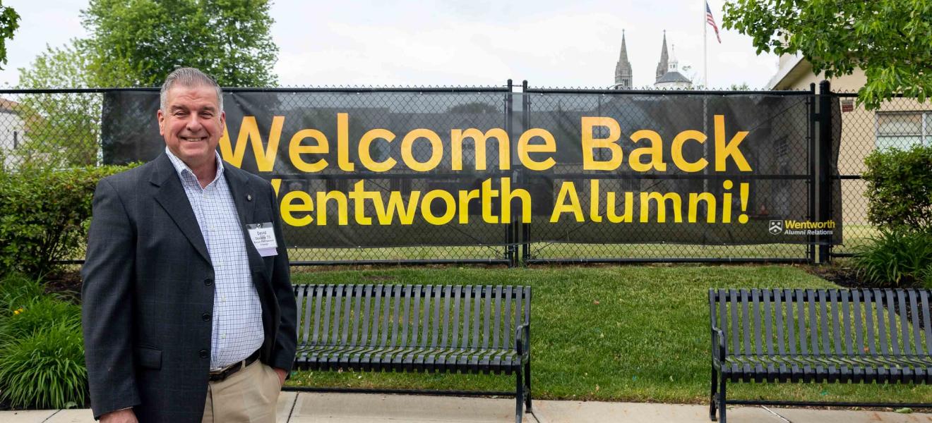 a man stands in front of a banner reading "alumni"