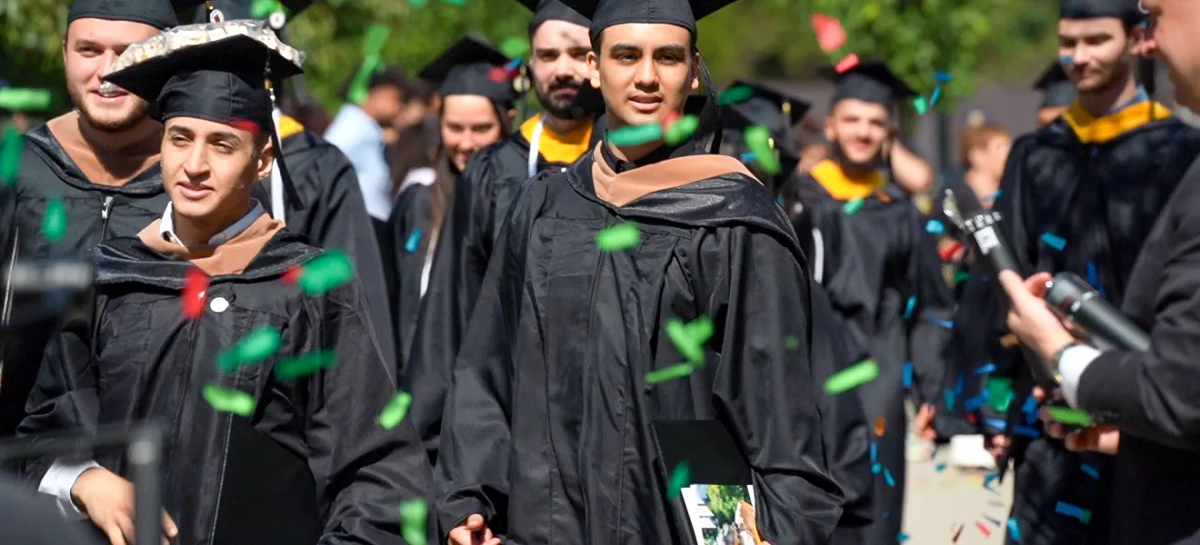 students wearing graduation robes walk through falling confetti 