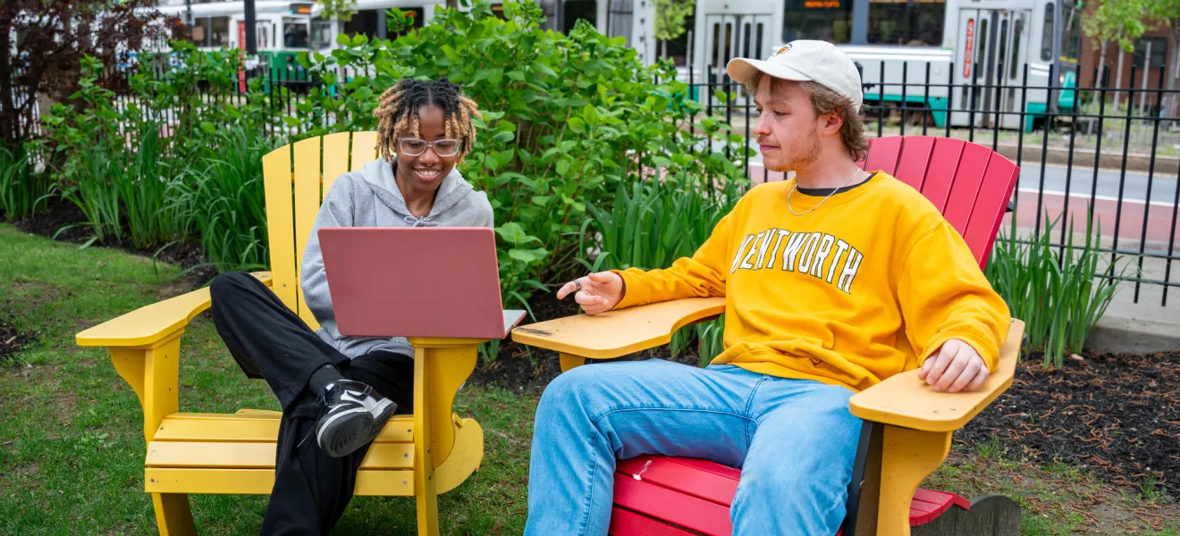 A Black female student and a white male wearing a Wentworth shirt sit together on the quad to study looking at a laptop. The Green Line T is in the background.