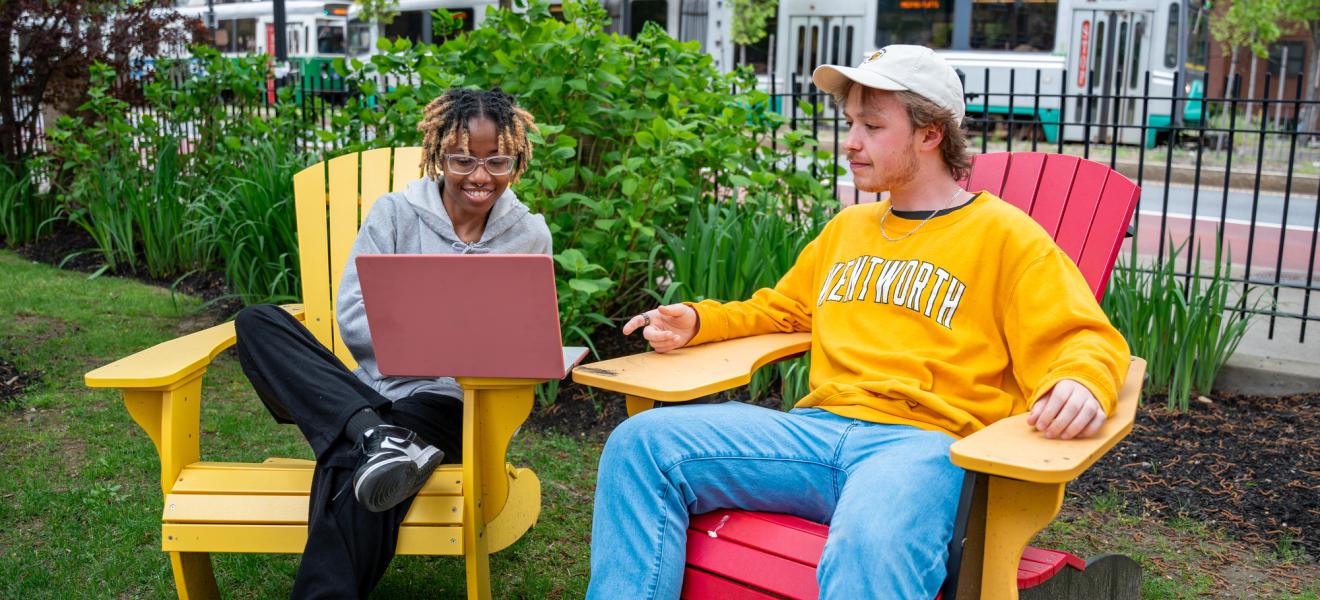 A Black female student and a white male wearing a Wentworth shirt sit together on the quad to study looking at a laptop. The Green Line T is in the background.