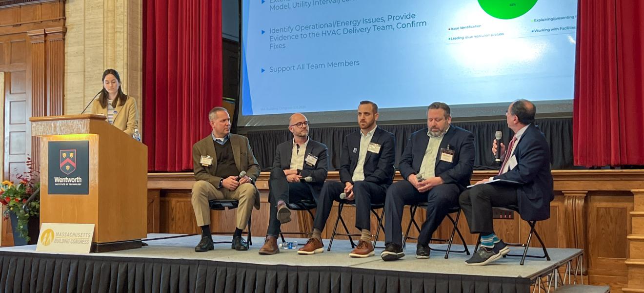 four people sit on a stage as part of a panel while a moderator stands at a podium