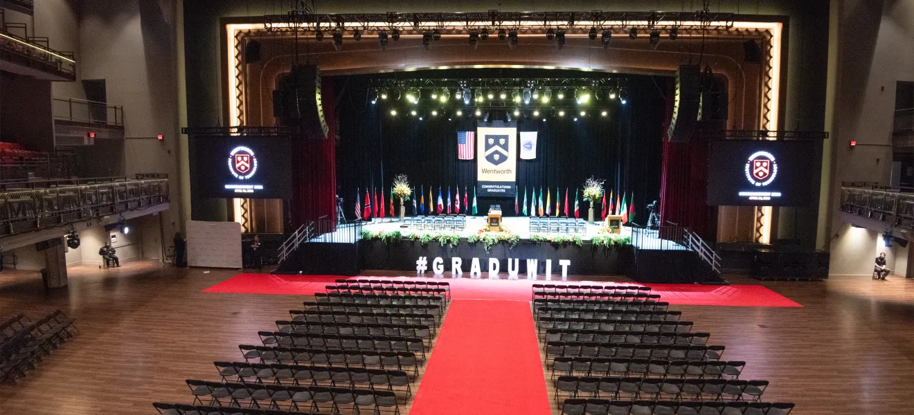 View of stage for commencement - red carpet flanked by black chairs for student seating, white cut out letters #graduwit, yellow, red and green florals decorating the black stage front, 26 international flags on poles representing the countries of international students, large Wentworth banner in gold and black with our school seal flanked by the US and Massachusetts state flags, 2 podiums and 35 black padded chairs on the stage for faculty and trustees