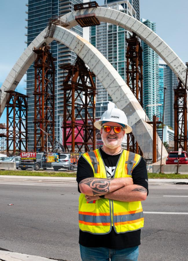 Jason Hatcher wearing a hardhat and safety vest stands in front a partially built bridge he is working on.